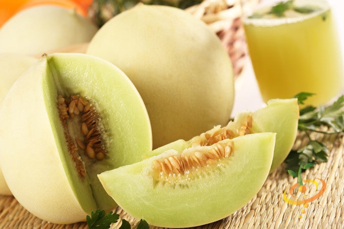 Honeydew melon with green flesh and seeds, whole and sliced on a table with a juice glass in the background